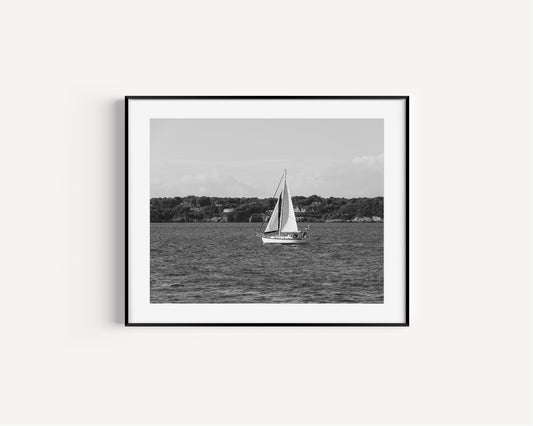 Horizontal black and white sailboat photo taken in Newport, Rhode Island, featuring a single sailboat on open water with a calm shoreline in the background. Ideal for coastal wall decor and lake house art.