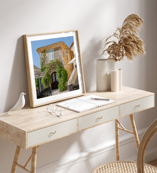 Square color print of a sunlit building in the village of Èze, featuring light blue shutters, ivy-covered stone walls, and a patio table—perfect for French country decor and Côte d’Azur inspired styling.