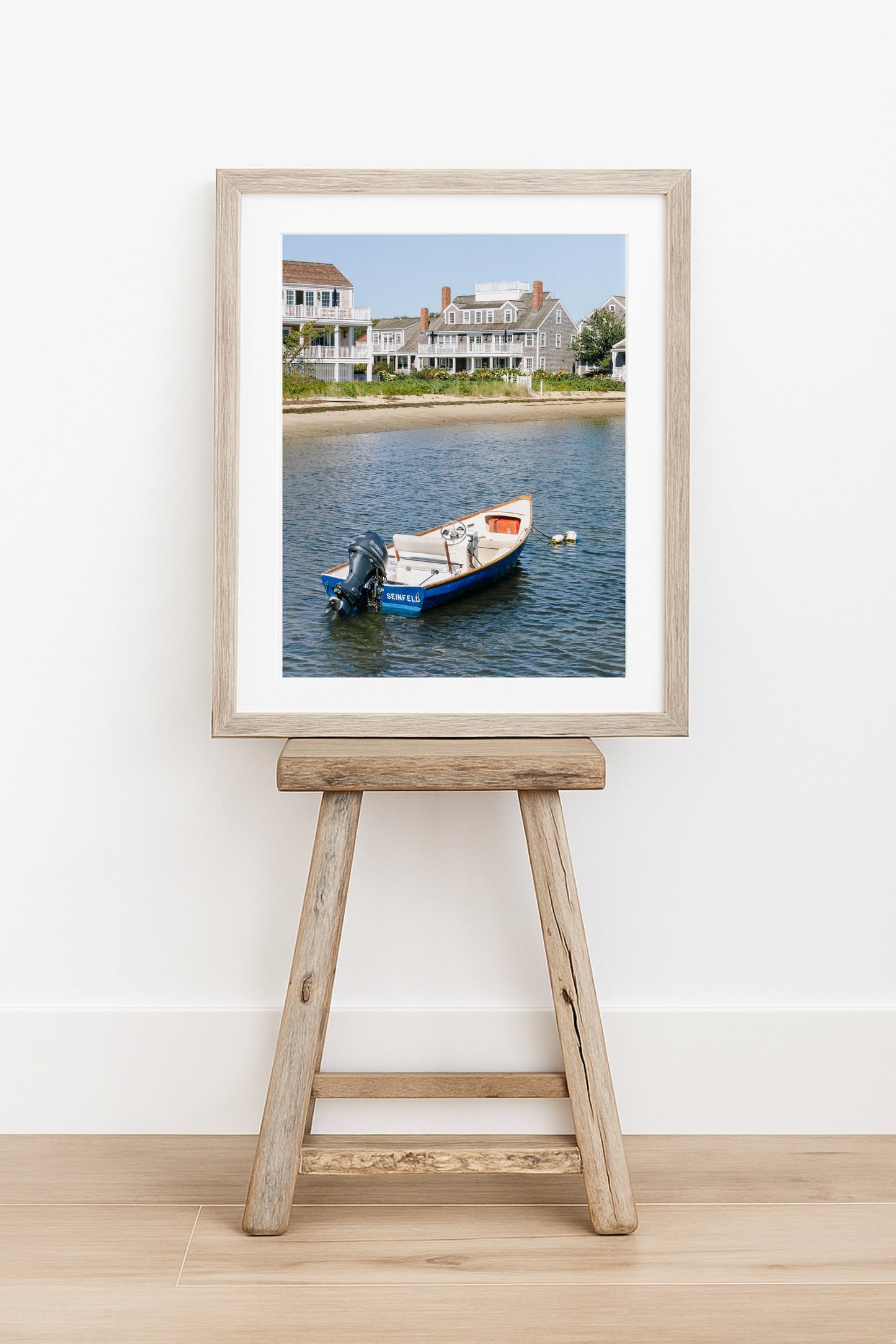 Vertical beach photography print of a small blue boat floating in Nantucket harbor, with Cape Cod-style houses and sandy shoreline in the background — perfect for nautical home decor and coastal wall art.