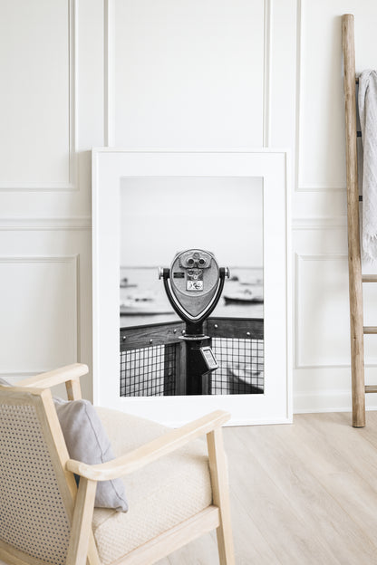 Black and white fine art photograph of a coin-operated viewfinder binoculars on Chatham Pier, Cape Cod — boats soft in the harbor background, wooden pier railing in foreground