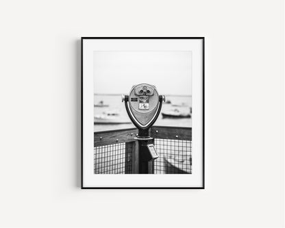 Black and white fine art photograph of a coin-operated viewfinder binoculars on Chatham Pier, Cape Cod — boats soft in the harbor background, wooden pier railing in foreground