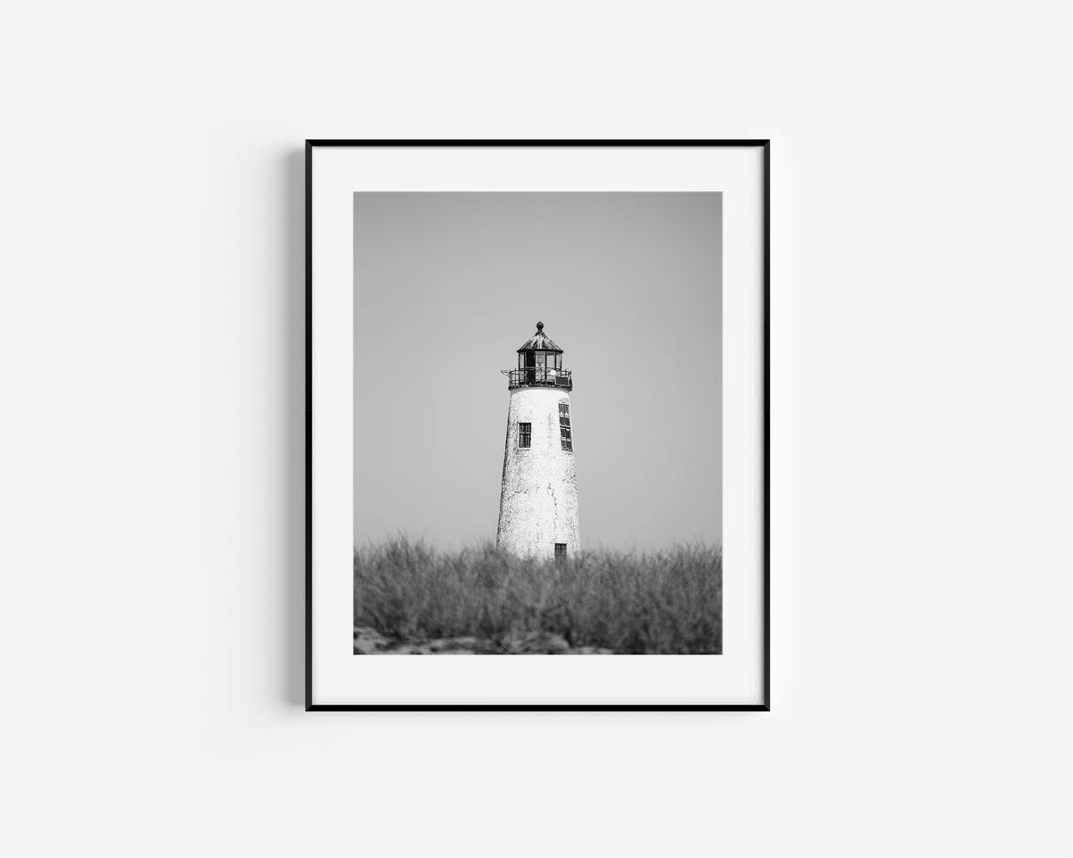 Black and white fine art photograph of Great Point Lighthouse and keeper's house on Nantucket Island — weathered white tower above dark dune grass against a flat grey sky, remote coastal wall art