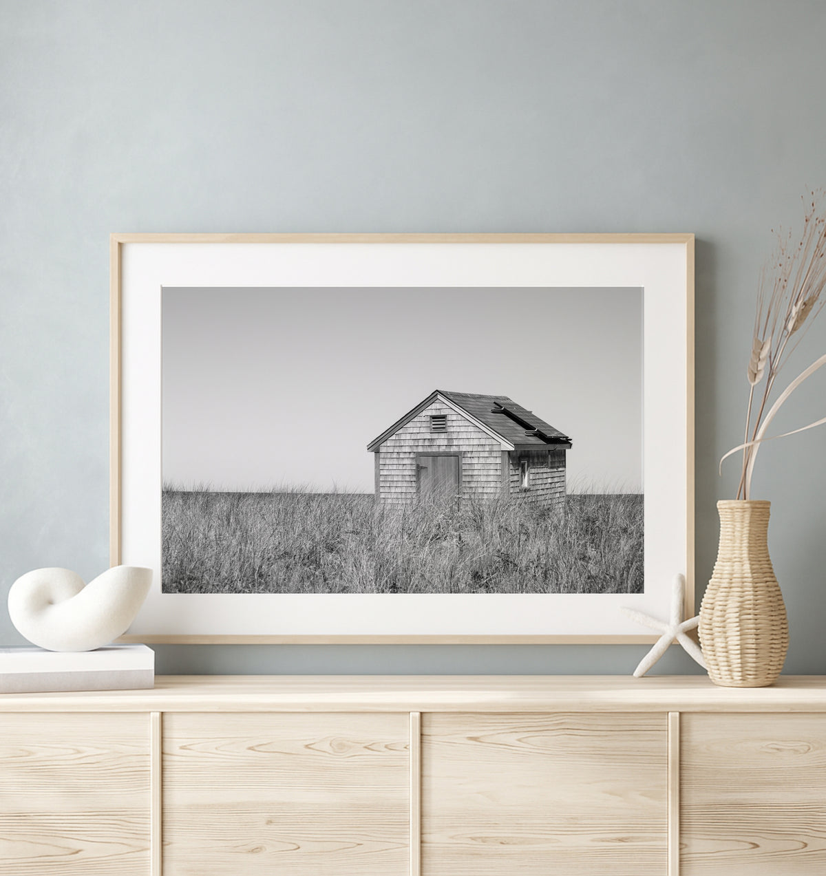 Black and white fine art photograph of a weathered cedar shingle beach cottage alone in wild dune grass on Nantucket Island — flat grey sky above, ocean just visible on the horizon