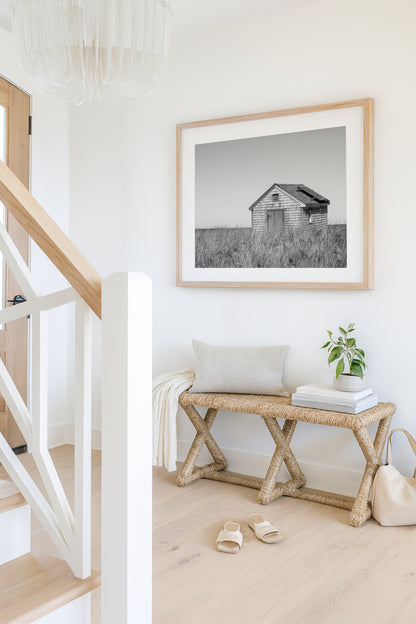 Black and white fine art photograph of a weathered cedar shingle beach cottage alone in wild dune grass on Nantucket Island — flat grey sky above, ocean just visible on the horizon