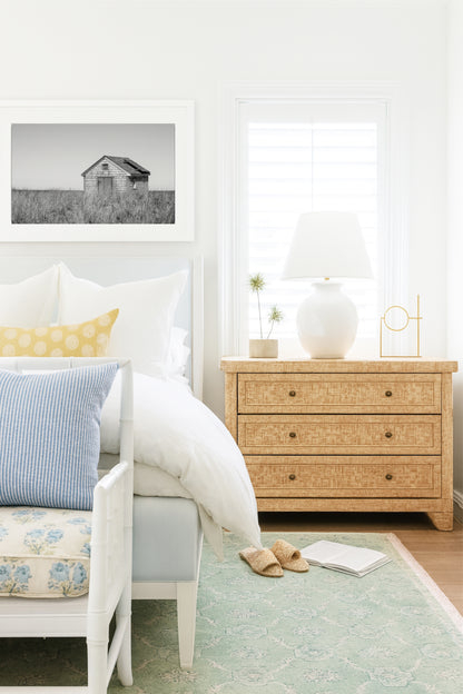 Black and white fine art photograph of a weathered cedar shingle beach cottage alone in wild dune grass on Nantucket Island — flat grey sky above, ocean just visible on the horizon