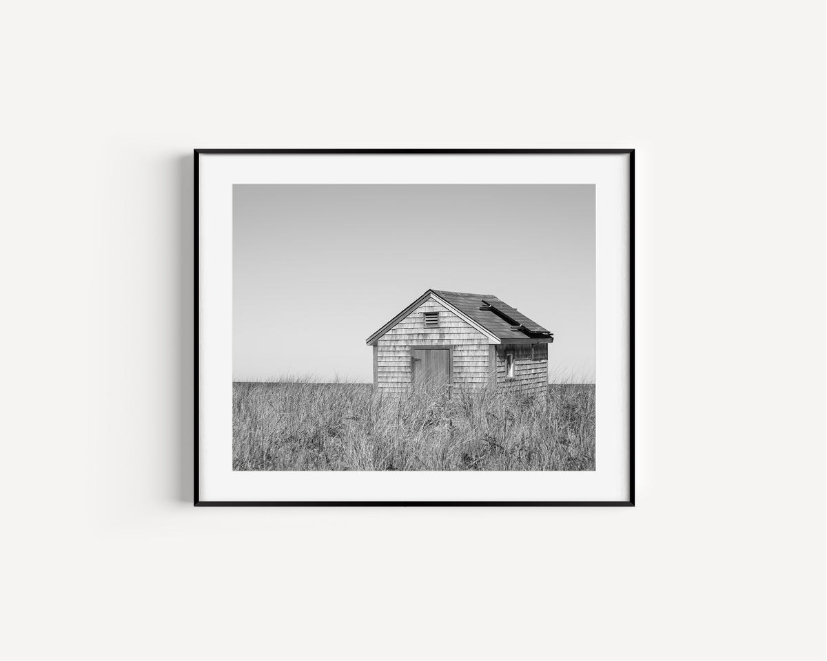 Black and white fine art photograph of a weathered cedar shingle beach cottage alone in wild dune grass on Nantucket Island — flat grey sky above, ocean just visible on the horizon
