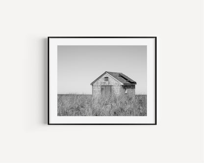 Black and white fine art photograph of a weathered cedar shingle beach cottage alone in wild dune grass on Nantucket Island — flat grey sky above, ocean just visible on the horizon