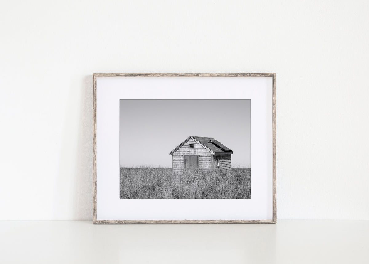 Black and white fine art photograph of a weathered cedar shingle beach cottage alone in wild dune grass on Nantucket Island — flat grey sky above, ocean just visible on the horizon