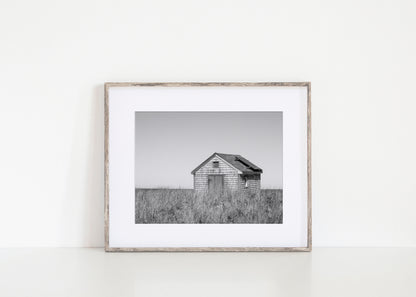 Black and white fine art photograph of a weathered cedar shingle beach cottage alone in wild dune grass on Nantucket Island — flat grey sky above, ocean just visible on the horizon