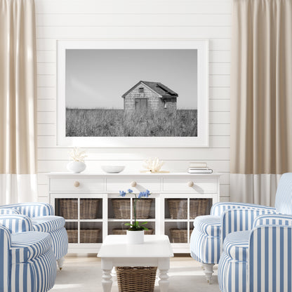 Black and white fine art photograph of a weathered cedar shingle beach cottage alone in wild dune grass on Nantucket Island — flat grey sky above, ocean just visible on the horizon