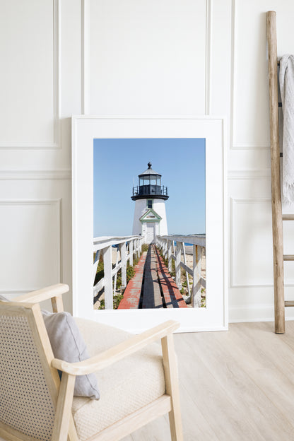 Vertical photograph of Brant Point Lighthouse in Nantucket, viewed straight down the weathered boardwalk with a bright blue sky and soft coastal light.