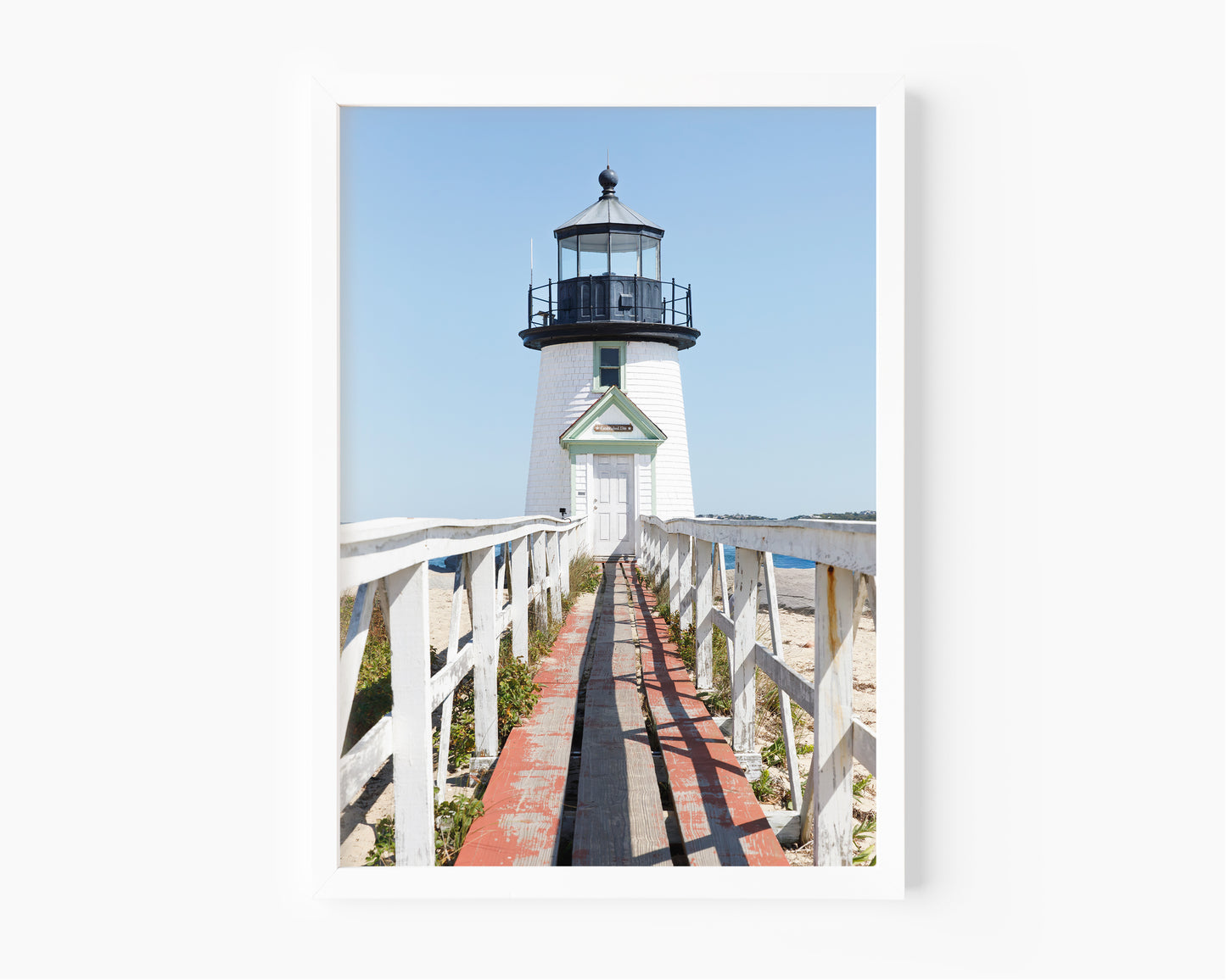 Vertical photograph of Brant Point Lighthouse in Nantucket, viewed straight down the weathered boardwalk with a bright blue sky and soft coastal light.