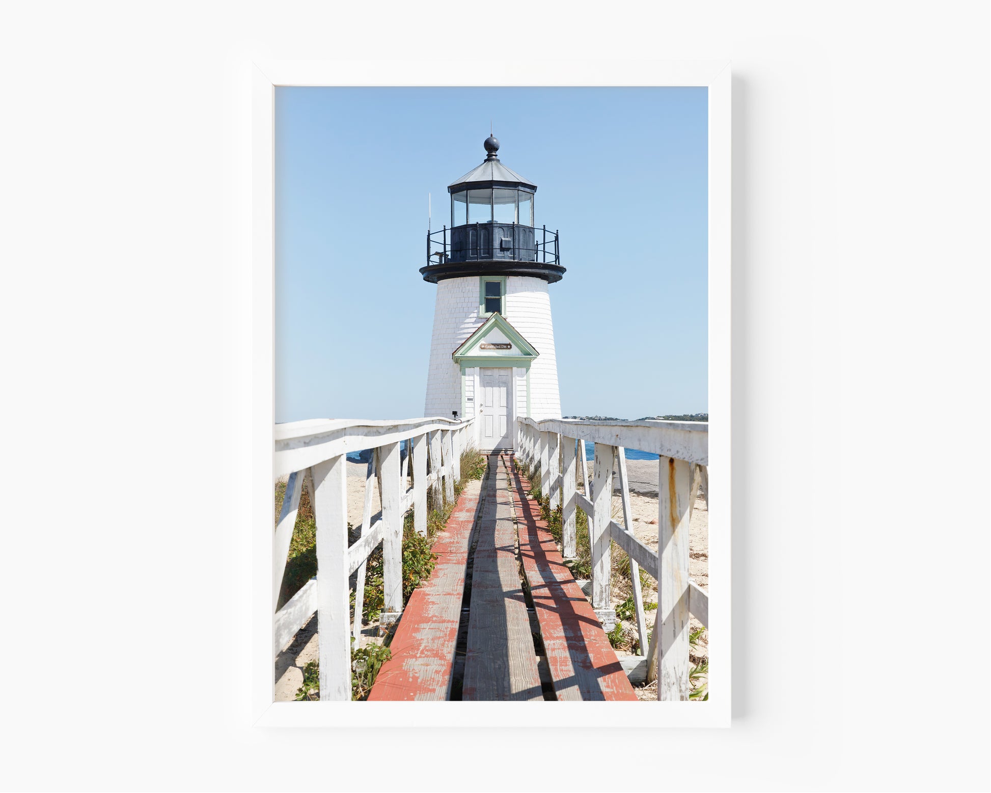 Vertical photograph of Brant Point Lighthouse in Nantucket, viewed straight down the weathered boardwalk with a bright blue sky and soft coastal light.