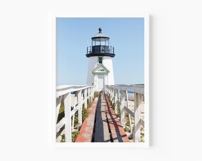 Vertical photograph of Brant Point Lighthouse in Nantucket, viewed straight down the weathered boardwalk with a bright blue sky and soft coastal light.