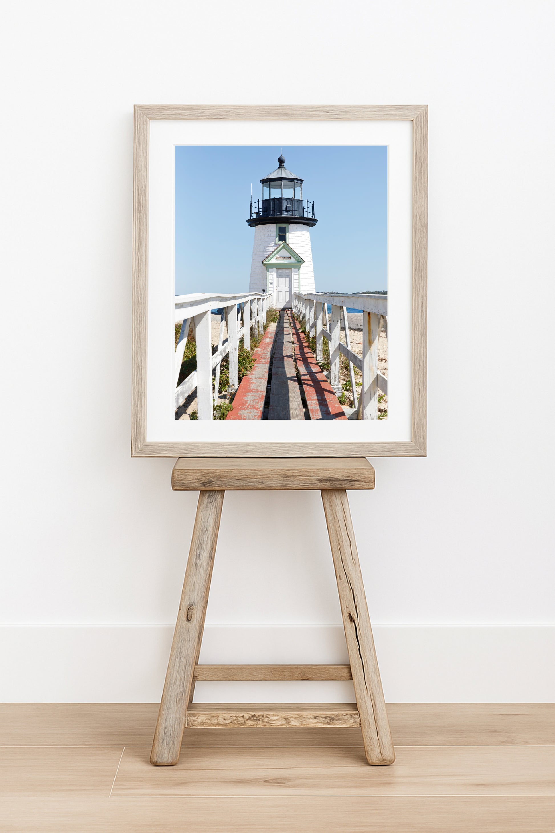 Vertical photograph of Brant Point Lighthouse in Nantucket, viewed straight down the weathered boardwalk with a bright blue sky and soft coastal light.