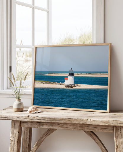 Color fine art photograph of Brant Point Lighthouse on Nantucket in horizontal format — white tower with red roof centered on a wide sandy spit surrounded by deep blue Nantucket Harbor