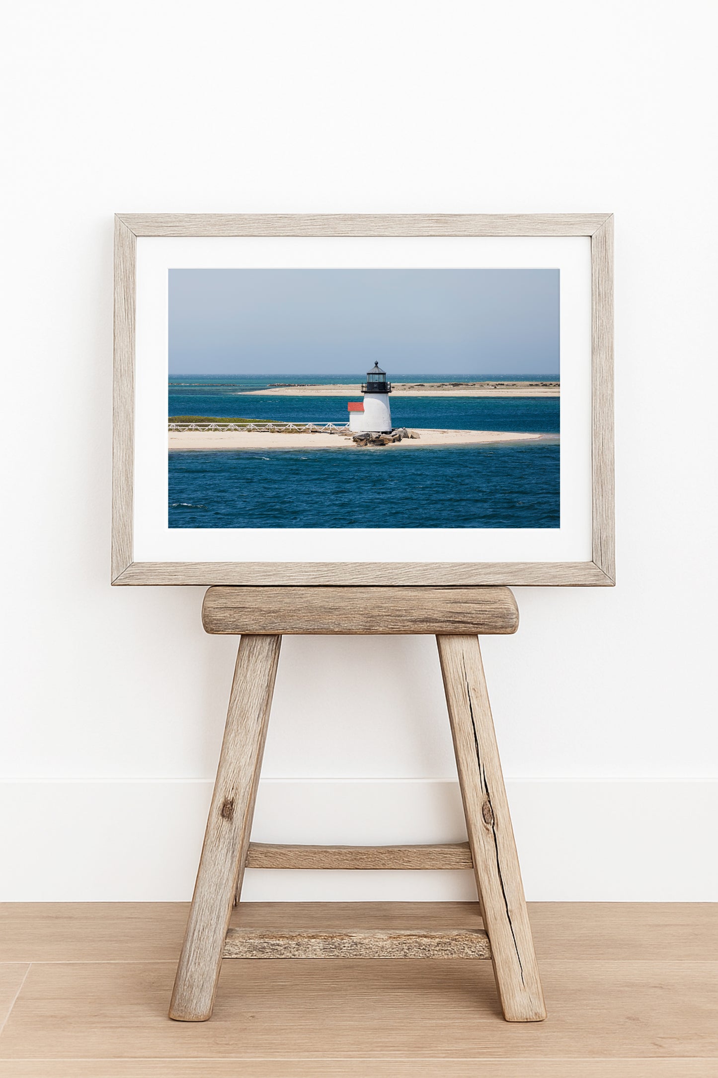 Color fine art photograph of Brant Point Lighthouse on Nantucket in horizontal format — white tower with red roof centered on a wide sandy spit surrounded by deep blue Nantucket Harbor