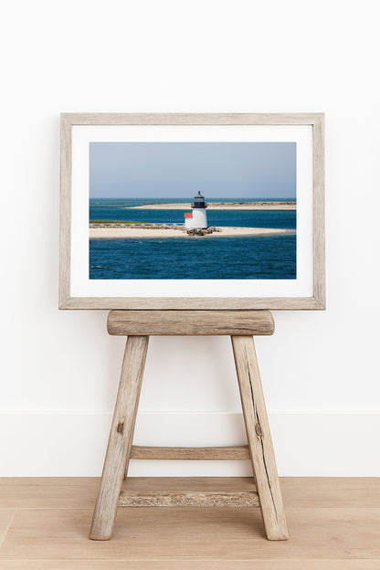 Color fine art photograph of Brant Point Lighthouse on Nantucket in horizontal format — white tower with red roof centered on a wide sandy spit surrounded by deep blue Nantucket Harbor