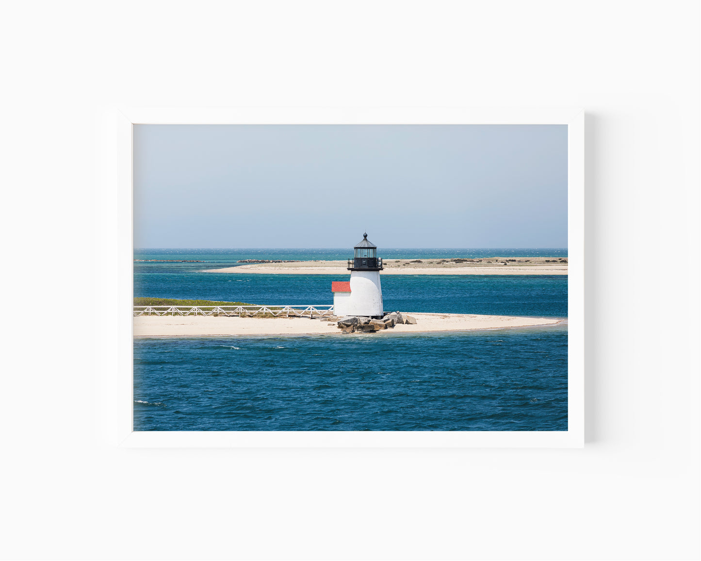 Color fine art photograph of Brant Point Lighthouse on Nantucket in horizontal format — white tower with red roof centered on a wide sandy spit surrounded by deep blue Nantucket Harbor