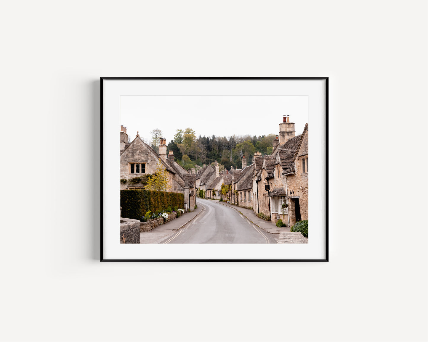 Color fine art photograph of Castle Combe village in the Cotswolds, England — empty road winding through honey-stone cottages with woodland rising behind the rooftops