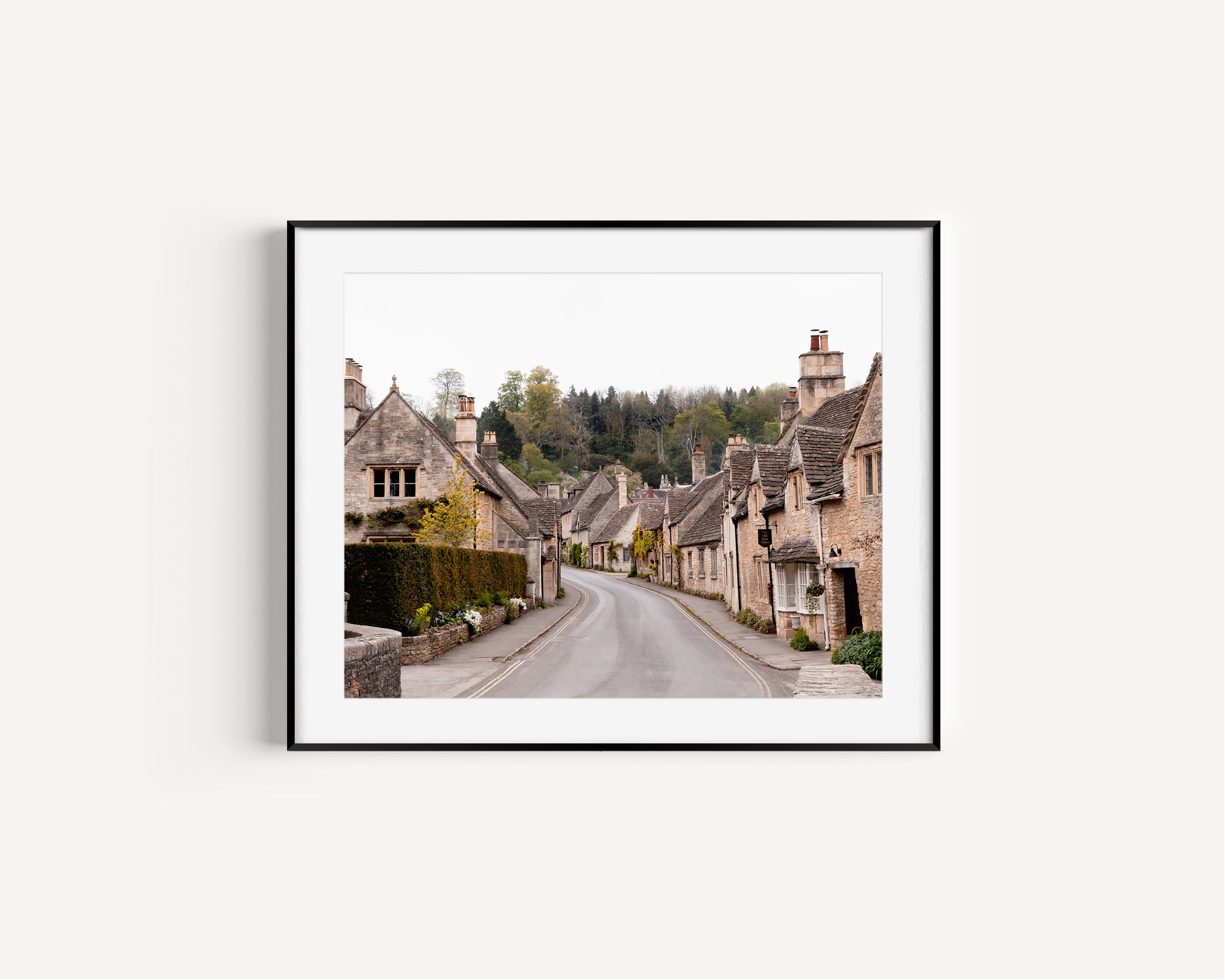 Color fine art photograph of Castle Combe village in the Cotswolds, England — empty road winding through honey-stone cottages with woodland rising behind the rooftops