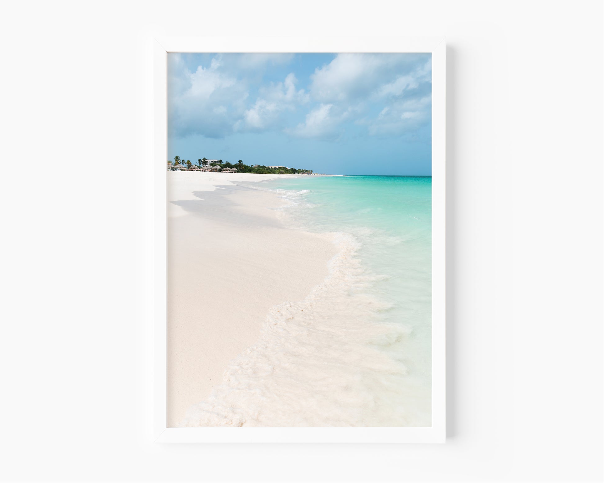 Color fine art photograph of Eagle Beach in Aruba — powder white sand, turquoise Caribbean water with a gentle wave at the shoreline, and palapa huts visible in the distance
