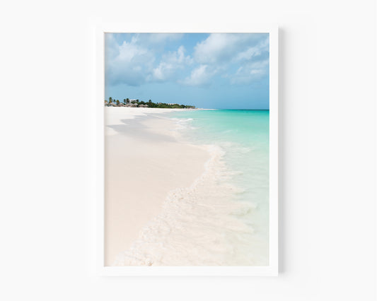 Color fine art photograph of Eagle Beach in Aruba — powder white sand, turquoise Caribbean water with a gentle wave at the shoreline, and palapa huts visible in the distance