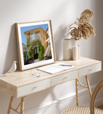 Square color print of a sunlit building in the village of Èze, featuring light blue shutters, ivy-covered stone walls, and a patio table—perfect for French country decor and Côte d’Azur inspired styling.