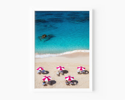 Beach scene with people under pink and white umbrellas, clear blue water, and white sand.