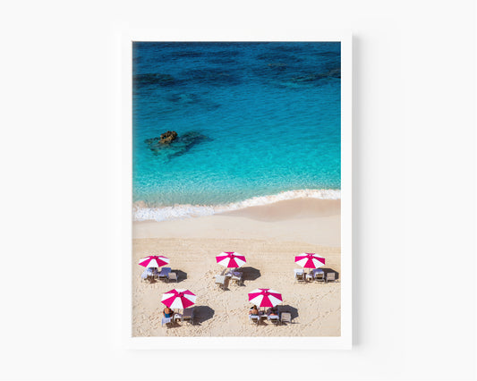 Beach scene with people under pink and white umbrellas, clear blue water, and white sand.