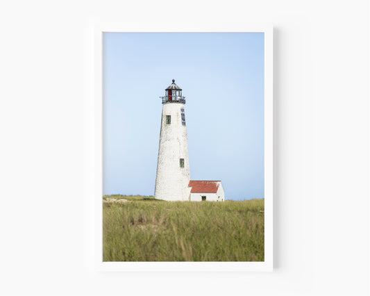 Color fine art photograph of Great Point Lighthouse at the northern tip of Nantucket Island — weathered white tower rising above green marsh grass with a small red-roofed keeper's house and pale blue sky