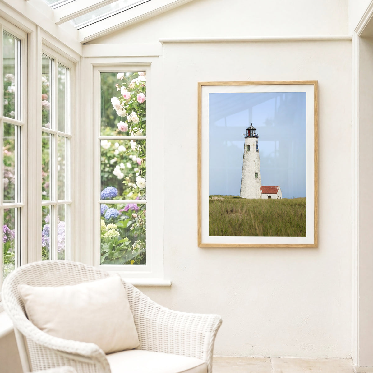 Color fine art photograph of Great Point Lighthouse at the northern tip of Nantucket Island — weathered white tower rising above green marsh grass with a small red-roofed keeper's house and pale blue sky 