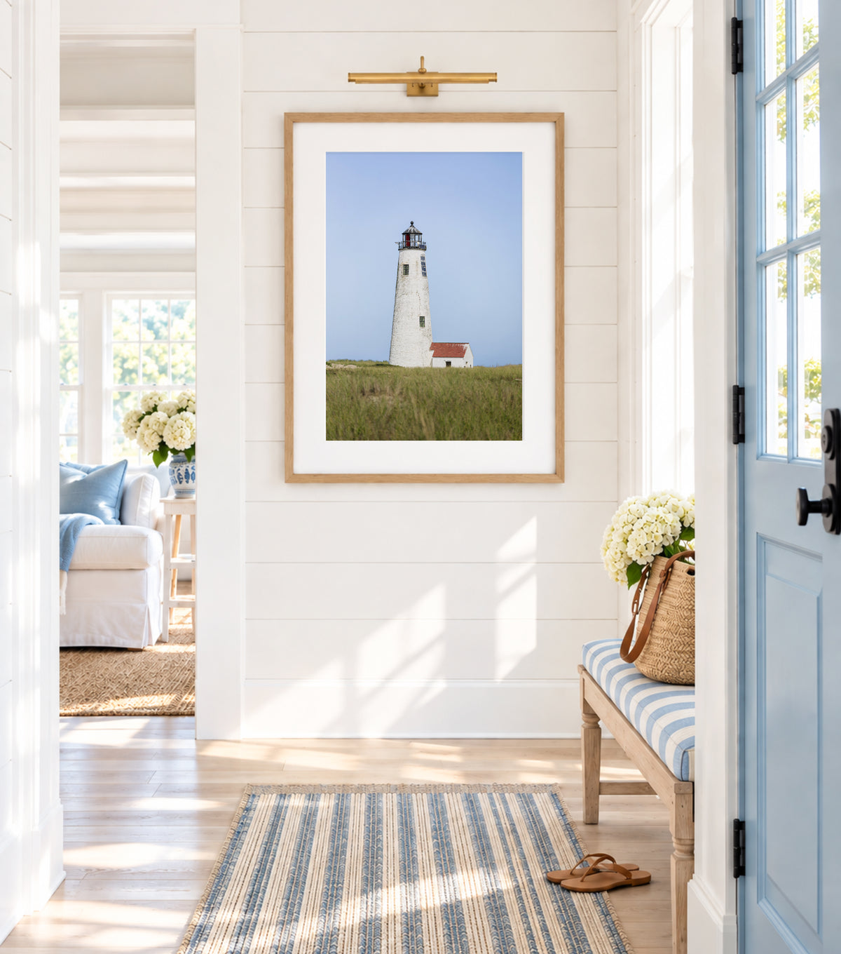 Color fine art photograph of Great Point Lighthouse at the northern tip of Nantucket Island — weathered white tower rising above green marsh grass with a small red-roofed keeper's house and pale blue sky