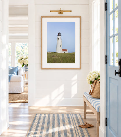 Color fine art photograph of Great Point Lighthouse at the northern tip of Nantucket Island — weathered white tower rising above green marsh grass with a small red-roofed keeper's house and pale blue sky