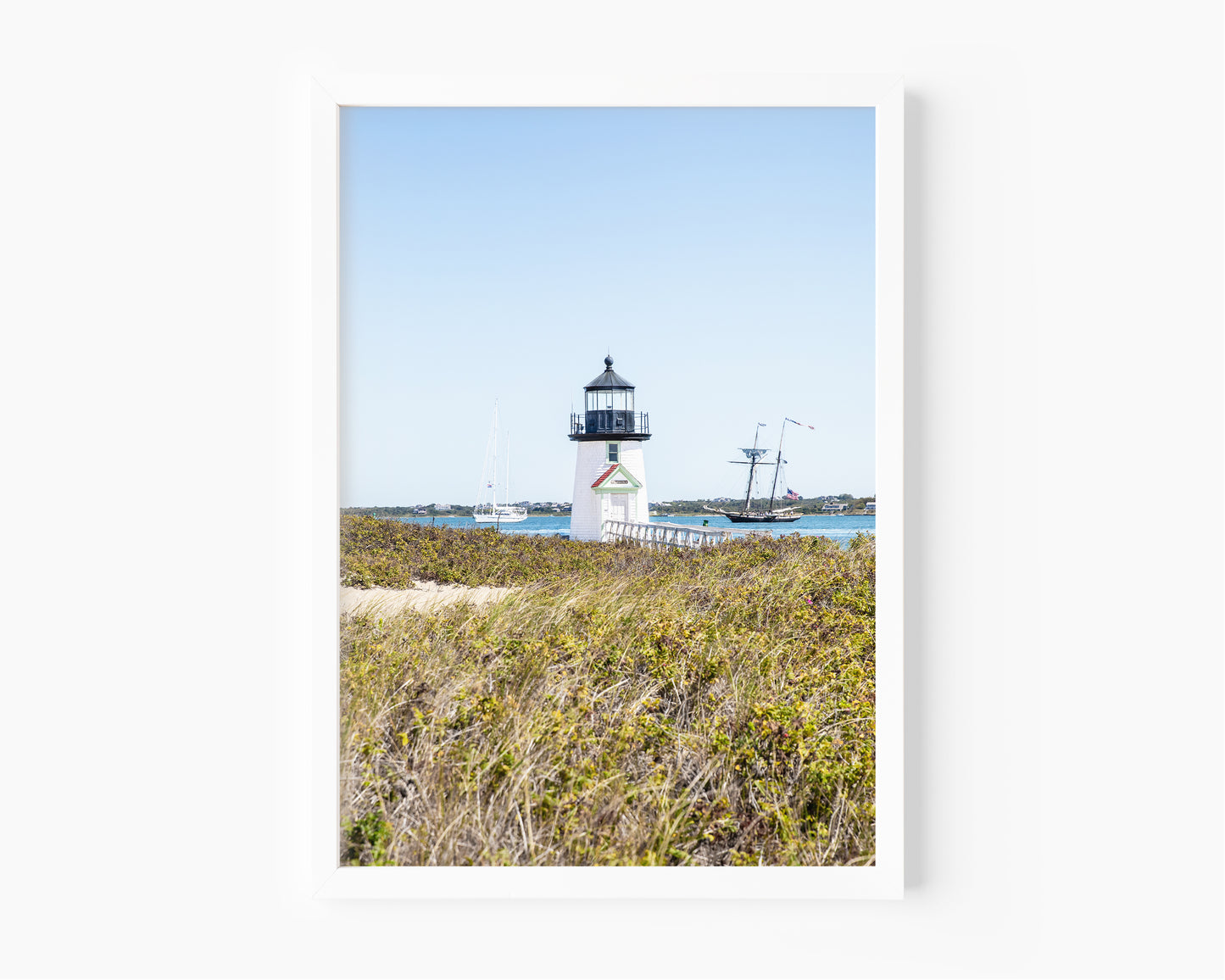 Brant Point Lighthouse stands at the edge of Nantucket Harbor, framed by dune grass with sailboats and a schooner in the background under a clear blue sky.