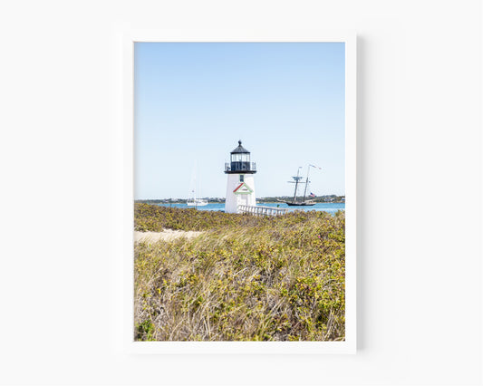 Brant Point Lighthouse stands at the edge of Nantucket Harbor, framed by dune grass with sailboats and a schooner in the background under a clear blue sky.