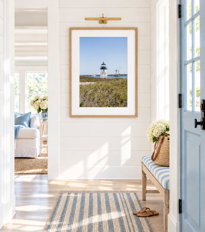 Brant Point Lighthouse stands at the edge of Nantucket Harbor, framed by dune grass with sailboats and a schooner in the background under a clear blue sky.