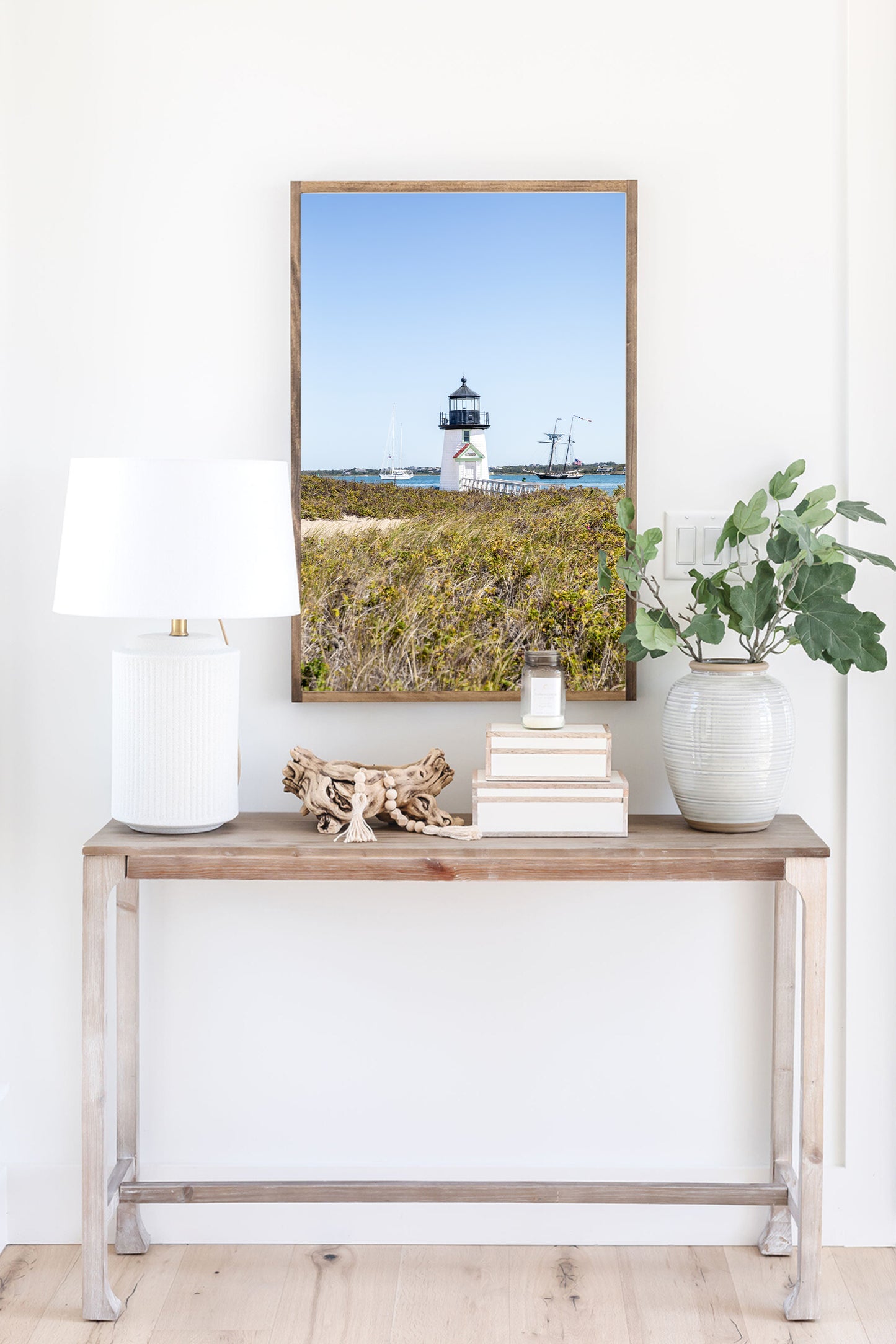 Brant Point Lighthouse stands at the edge of Nantucket Harbor, framed by dune grass with sailboats and a schooner in the background under a clear blue sky.