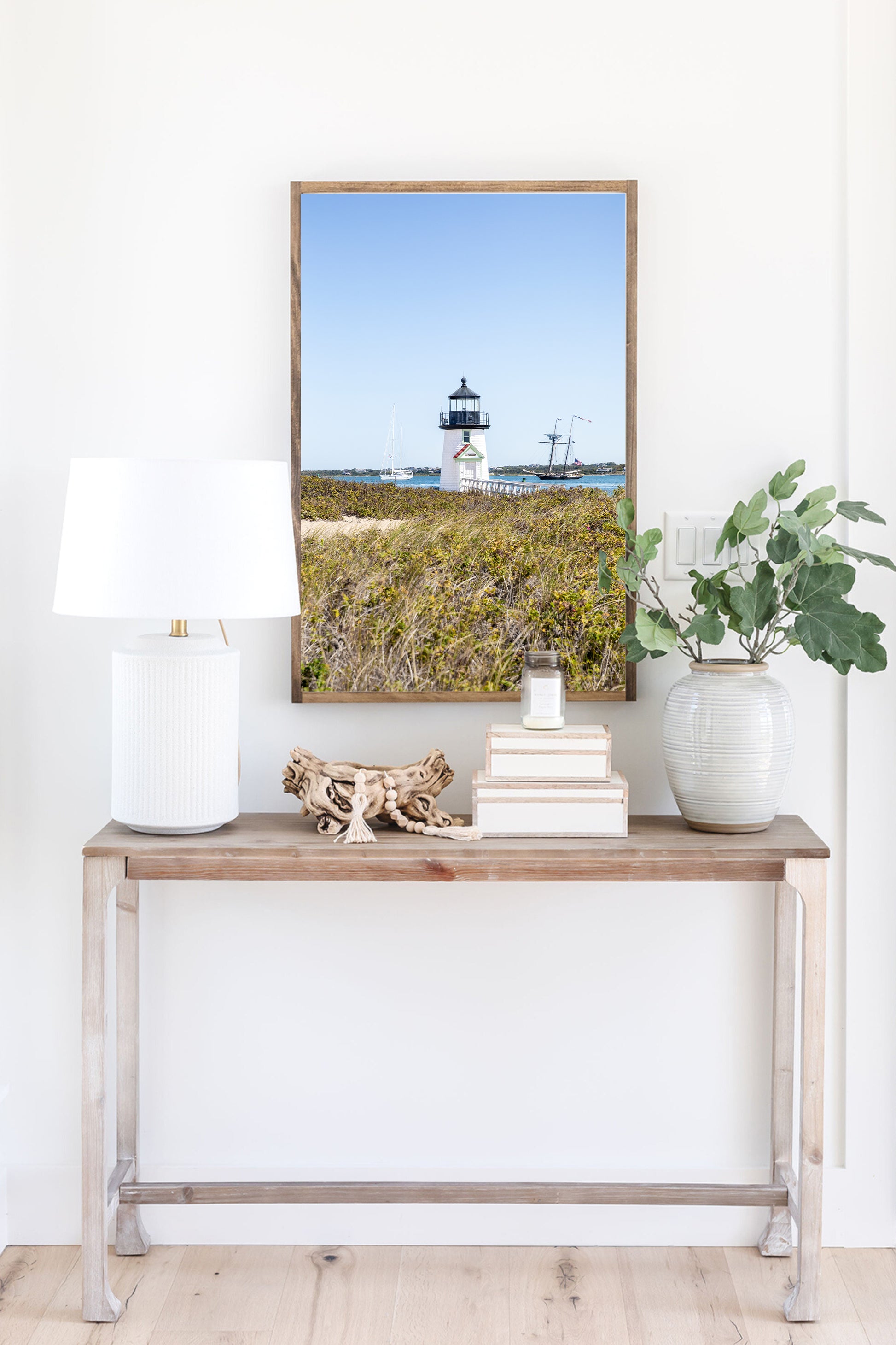 Brant Point Lighthouse stands at the edge of Nantucket Harbor, framed by dune grass with sailboats and a schooner in the background under a clear blue sky.