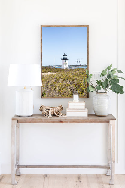 Brant Point Lighthouse stands at the edge of Nantucket Harbor, framed by dune grass with sailboats and a schooner in the background under a clear blue sky.