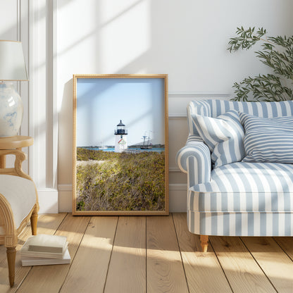 Brant Point Lighthouse stands at the edge of Nantucket Harbor, framed by dune grass with sailboats and a schooner in the background under a clear blue sky.