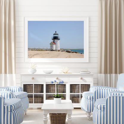 Color fine art photograph of Brant Point Lighthouse on Nantucket shot from the beach, white tower with red-roofed keeper's entrance, white fence boardwalk on the left and harbor water on both sides