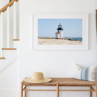 Color fine art photograph of Brant Point Lighthouse on Nantucket shot from the beach, white tower with red-roofed keeper's entrance, white fence boardwalk on the left and harbor water on both sides