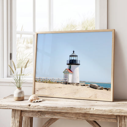 Color fine art photograph of Brant Point Lighthouse on Nantucket shot from the beach, white tower with red-roofed keeper's entrance, white fence boardwalk on the left and harbor water on both sides