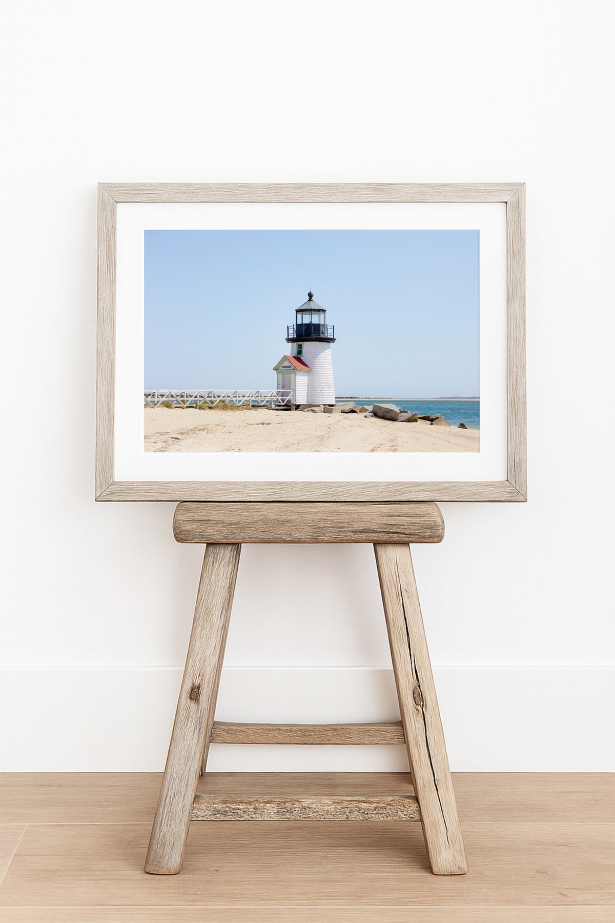 Color fine art photograph of Brant Point Lighthouse on Nantucket shot from the beach, white tower with red-roofed keeper's entrance, white fence boardwalk on the left and harbor water on both sides