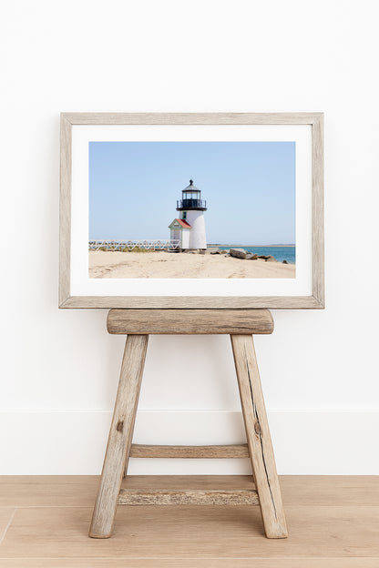 Color fine art photograph of Brant Point Lighthouse on Nantucket shot from the beach, white tower with red-roofed keeper's entrance, white fence boardwalk on the left and harbor water on both sides