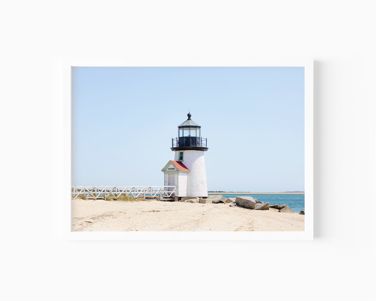 Color fine art photograph of Brant Point Lighthouse on Nantucket shot from the beach, white tower with red-roofed keeper's entrance, white fence boardwalk on the left and harbor water on both sides