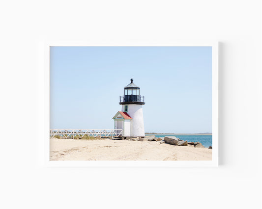 Color fine art photograph of Brant Point Lighthouse on Nantucket shot from the beach, white tower with red-roofed keeper's entrance, white fence boardwalk on the left and harbor water on both sides