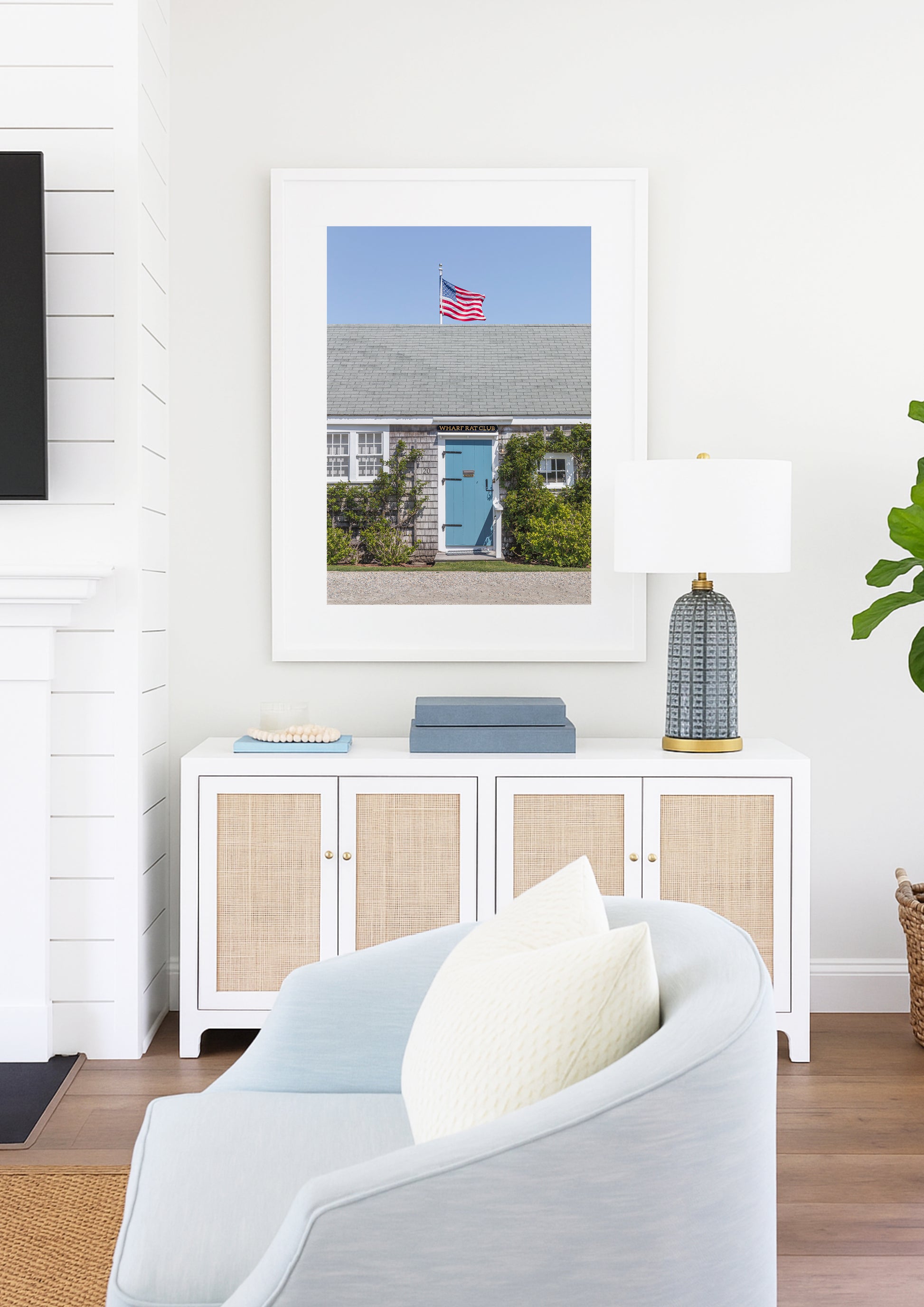 Color fine art photograph of the Wharf Rat Club on Nantucket — weathered grey shingle cottage with a powder blue door, climbing vines, and an American flag flying above the roofline against a blue summer sky