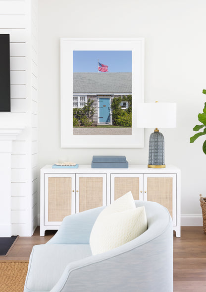 Color fine art photograph of the Wharf Rat Club on Nantucket — weathered grey shingle cottage with a powder blue door, climbing vines, and an American flag flying above the roofline against a blue summer sky